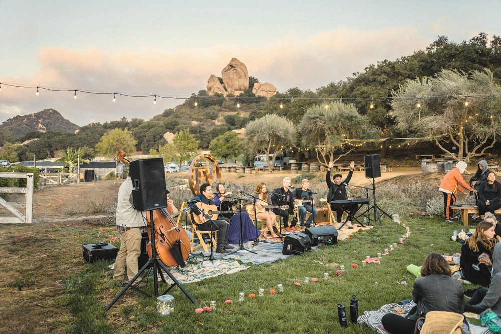 A full band on a lawn with sound and lights, guests seated on the grass below a rocky peak