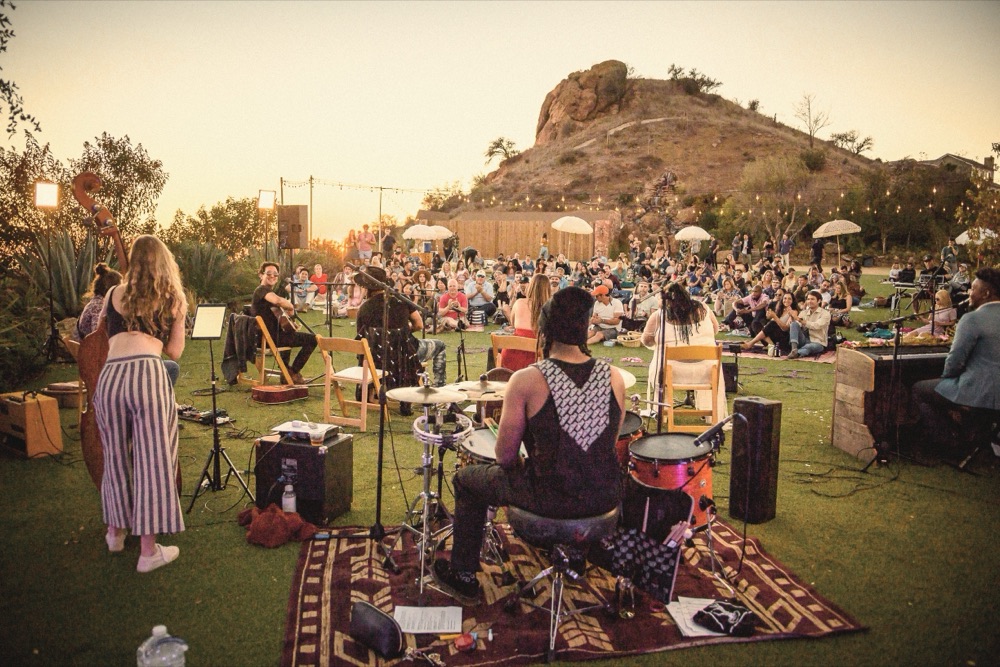 A band playing on a rug on the lawn at sunset, the audience seated on the grass